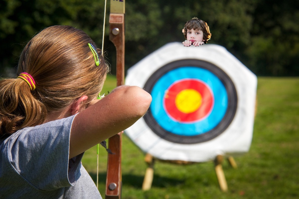 A modern William Tell's son, an innocent young boy peeks out above an archery practice target with an arrow trained on it