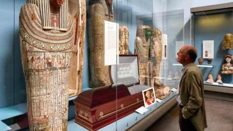 A man looks at a modern wooden coffin with a picture of Queen Elizabeth next to it that is behind the glass of a museum exhibit about Egyptian mummies