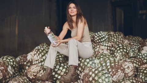 Kendall Jenner sits on top of a pile of agave, wearing an entirely beige outfit with brown boots, holding a bottle of 818 tequila, and staring in the general direction of the camera with an expression of pride