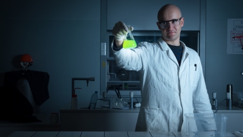 A bald man wearing a white coat, gloves, and goggles is standing in a dark lab and holding up a glowing green erlenmeyer flask