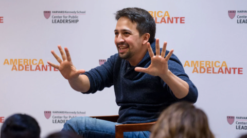 Lin-Manuel Miranda doing Jazzhands in front of a backdrop with the HKS logo