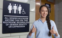 A girl with thumbs up outside a gender neutral bathroom