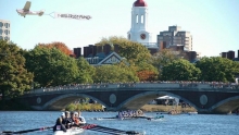Two crews row under weeks footbridge as spectators look on. A small plane tows a banner reading "1-800-Trust Fund."
