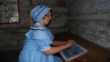 pioneer girl in blue bonnet and frilly dress writes on a slate tablet with chalk